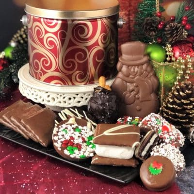 A holiday tin in a festive design surrounded by gourmet chocolate cookies and treats nestles among Christmas ornaments.